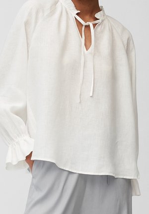 Witte losse blouse met lange mouwen, gerimpelde kraag en manchetten, kraag met strik, gedragen met lichtgrijze broek, hand in de zak.