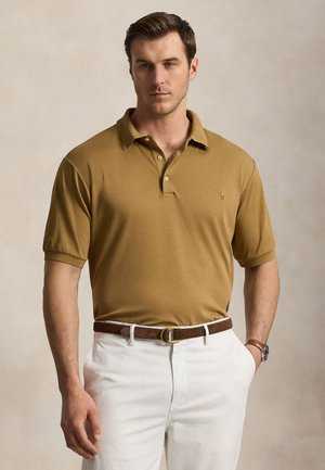 Mand iført en sennepsfarvet polo-shirt indbundet i hvide bukser med brunt bælte, stående mod en ensfarvet lys baggrund med den ene hånd i lommen.