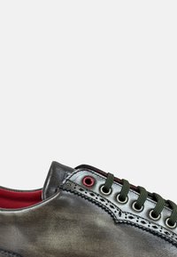 Zapato de cuero gris con una textura suave, cordones verdes, ojales rojos y costuras decorativas a lo largo del borde. El interior presenta un forro rojo.