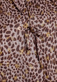 Tessuto con stampa leopardata nelle tonalità di marrone e oro, caratterizzato da una consistenza morbida e un dettaglio annodato al collo. Pattern a righe verticali visibile.