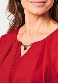 Rode blouse met een V-hals ontwerp, voorzien van een kettingaccent en een ronde hanger bij de halslijn, gemaakt van een gestructureerde, lichtgewicht stof.