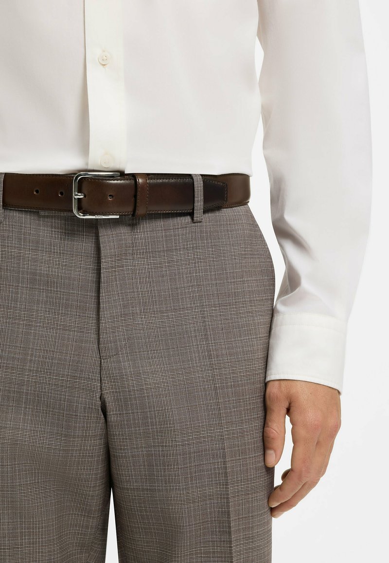 Homme portant une chemise blanche rentrée dans un pantalon à carreaux gris avec une ceinture en cuir marron foncé et une boucle argentée, main le long du corps.