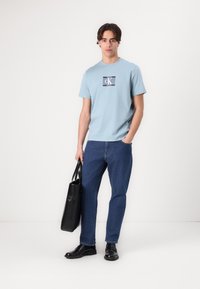 Lys blå bomulls T-skjorte med et rektangulært logo, sammen med medium vaskede dongeribukser. Modellen holder en svart skinn totebag og har på seg svarte sko.
