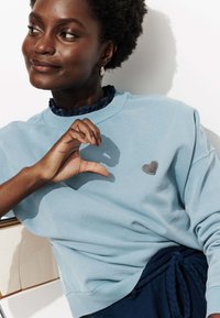 Sweatshirt bleu clair en coton avec un patch en forme de cœur texturé sur le côté gauche. Présente un col rond côtelé et une coupe décontractée.