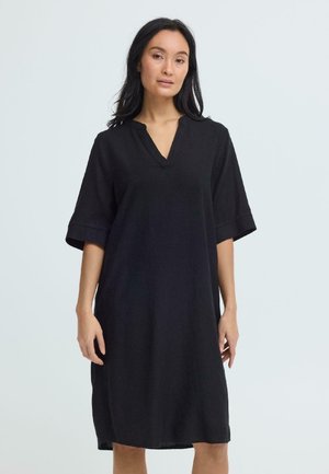 Kobieta stojąca w luźnej, czarnej sukience do kolan z krótkimi rękawami i dekoltem w serek na jednolitym jasnym tle.