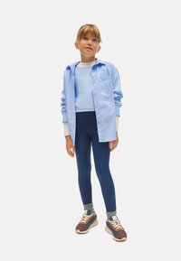 Camisa de botones a rayas azul claro sobre una blusa azul claro, combinada con leggings azul oscuro y zapatillas multicolor. Atuendo casual en capas.