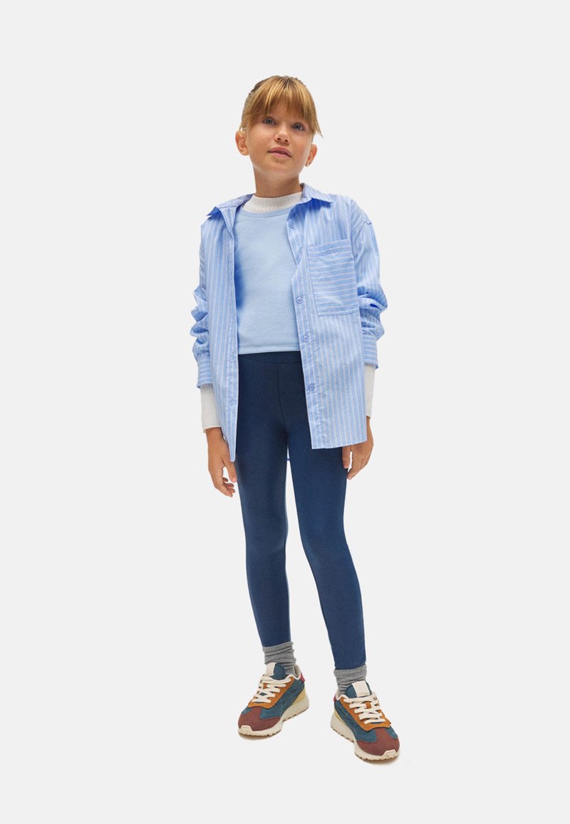 Camisa de botones a rayas azul claro sobre una blusa azul claro, combinada con leggings azul oscuro y zapatillas multicolor. Atuendo casual en capas.