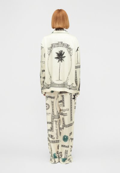 Άτομο με κοντά κόκκινα μαλλιά, ντυμένο με άνετο κρεμ ρούχο τυπωμένο με μαύρα και πράσινα σχέδια φοίνικα και διακοσμητικά πλαίσια, όρθιο με την πλάτη γυρισμένη.