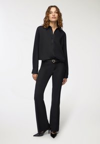 Camicia nera con bottoni e maniche lunghe, abbinata a pantaloni neri aderenti. Una cintura nera presenta una fibbia dorata a forma di cuore. Tessuto elegante.