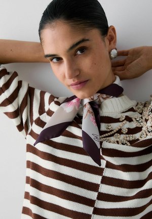 Donna con capelli scuri raccolti dietro indossa un maglione a righe marrone e bianco con ricami floreali, orecchini bianchi vistosi e una sciarpa di seta floreale al collo.