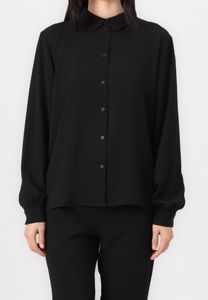 Personne portant un chemisier noir à manches longues boutonné avec un col et un pantalon noir assorti sur un fond uni.