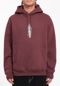 Sudadera color marrón oscuro hecha de tejido suave, con un bolsillo canguro frontal y un diseño gráfico de una figura en posición de pie.