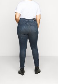 Jeans skinny azul oscuro con bolsillos traseros y cintura alta, combinados con una camiseta blanca y botines negros de tacón.