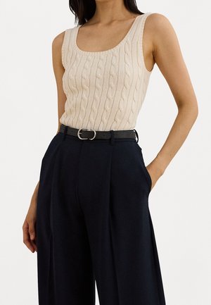 Vrouw die een crèmekleurige kabelgebreide mouwloze top draagt, gestopt in een zwarte, hooggesneden, wijde broek met een zwarte riem.