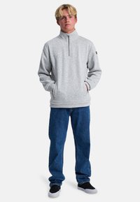 Sudadera de forro polar gris con cuello de media cremallera, que presenta un patrón texturizado y bolsillos laterales, combinada con jeans de mezclilla azules y zapatos negros.