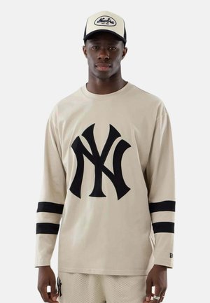 Giovane uomo che indossa una camicia beige a maniche lunghe con grande logo nero NY, pantaloncini in rete beige abbinati e cappellino beige New Era, in posizione neutra.