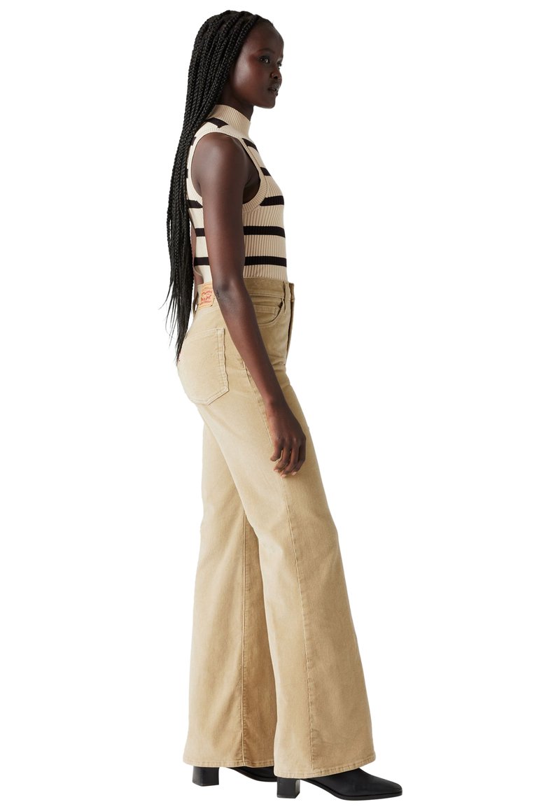 Jean large beige taille haute, associé à un haut sans manches rayé crème et marron. Texture lisse avec des coutures visibles.