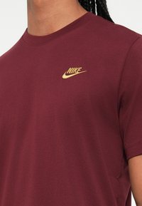 Osoba na sobě má vínové tričko Nike s malým zlatým logem Nike vyšitým na levé hrudi, s krátkým střihem na krku a rameni.