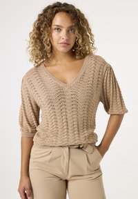 Beige gestrickter Pullover mit V-Ausschnitt, kurzen Puffärmel und einem strukturierten Muster. Kombiniert mit hellbeigen, hochgeschnittenen Hosen.