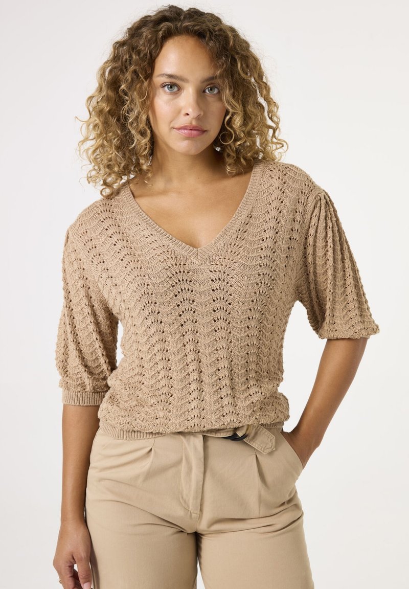 Beige gestrickter Pullover mit V-Ausschnitt, kurzen Puffärmel und einem strukturierten Muster. Kombiniert mit hellbeigen, hochgeschnittenen Hosen.