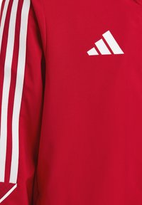 Rode sportjack met een witte driebaanontwerp op de schouder, een gladde stoffen textuur en een wit Adidas-logo op de borst.