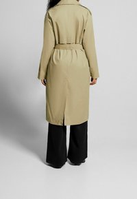Béžový trench coat s páskem v pase, dlouhými rukávy a rozparkem na zadní straně. Kombinovaný s širokými černými kalhotami, zobrazený zezadu na neutrálním pozadí.