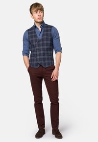 Granatowa kamizelka w kratę nałożona na niebieską koszulę, w zestawieniu z brązowymi spodniami i ciemnymi butami. Kamizelka ma podwójne zapięcie i kieszenie.