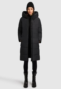 Čierna puffová bunda s kapucňou, s prešívaným dizajnom a bočnými zipsovými vreckami. Skombinovaná s čiernymi legínami a čižmami po kolená.