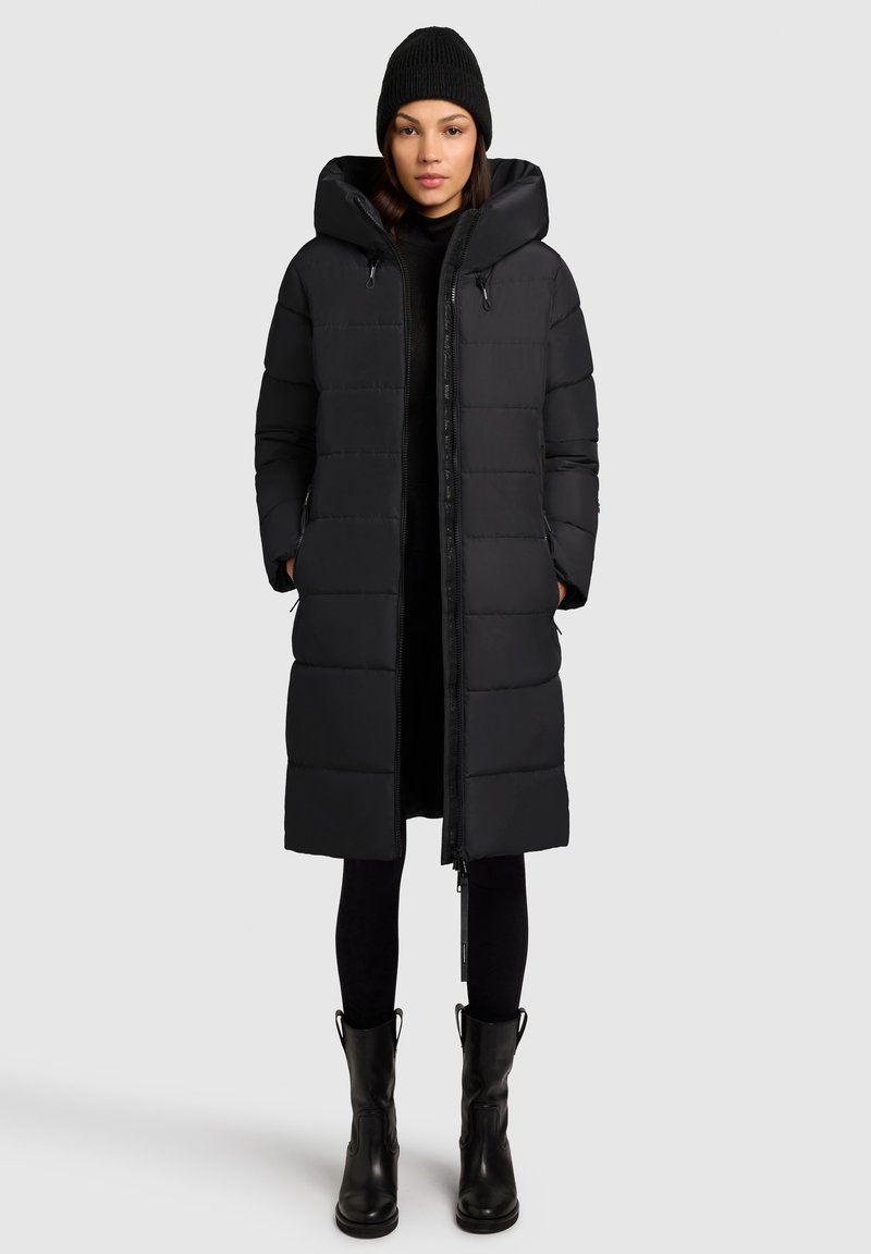 Čierna puffová bunda s kapucňou, s prešívaným dizajnom a bočnými zipsovými vreckami. Skombinovaná s čiernymi legínami a čižmami po kolená.