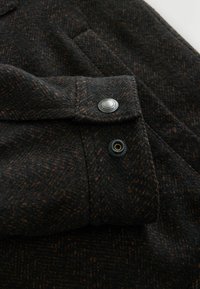 Tissu en laine sombre avec des taches brunes et un gros plan sur une manchette présentant un bouton-pression métallique marqué « Premium Quality ».