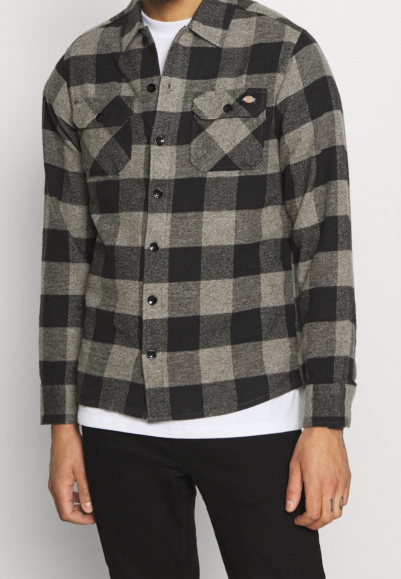 Camicia di flanella a scacchi nera e grigia con maniche lunghe, chiusura con bottoni e due tasche sul petto. Il tessuto ha un aspetto caldo e texture.