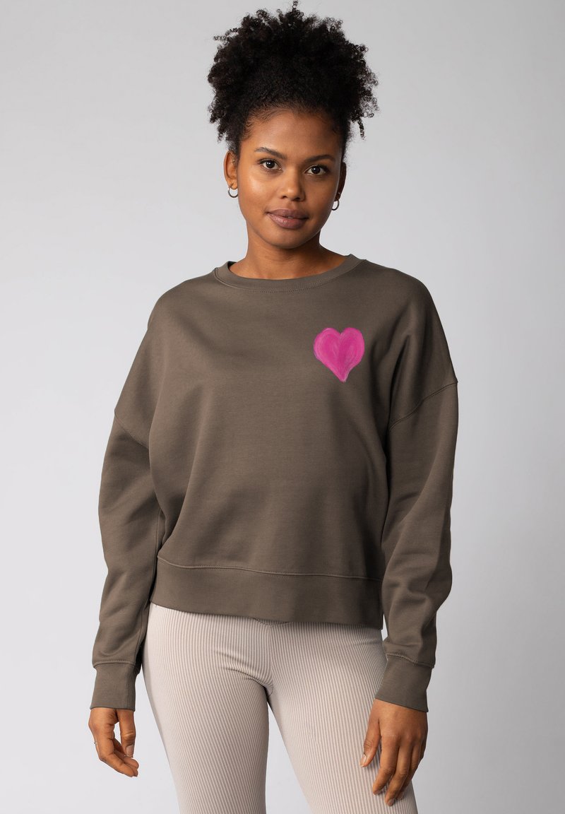 Bruine cropped sweatshirt met een roze hartontwerp op de linkerkant van de borst, gecombineerd met lichte geribde leggings. Zacht materiaal en een casual fit.