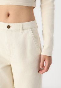 Beige Flatfront-Jeans mit strukturierter Oberfläche, ausgestattet mit seitlichen Taschen und einem Metallknopf-Akzent, kombiniert mit einem gerippten Oberteil.