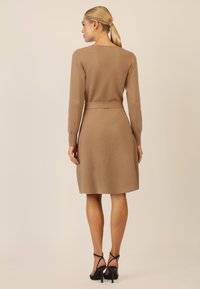 Strickkleid in hellbraun, mit langen Ärmeln, tailliertem Schnitt und schwingendem Rock, kombiniert mit schwarzen Riemchenschuhen.