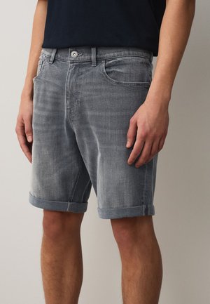 Man draagt omgeslagen grijze denimshorts en een donker shirt, staand tegen een effen lichte achtergrond.
