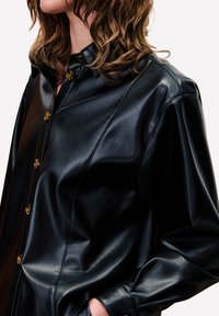 Zwarte leren shirt met een klassieke kraag, voorzien van gouden knopen en een relaxte pasvorm. De textuur lijkt glad en glanzend, wat het materiaal benadrukt.