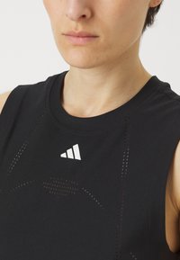Crna bezrukavna atletska majica s prozračnim mrežastim detaljima i bijelim logotipom na prsima; izrađena od lagane i rastezljive tkanine.