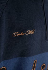 Granatowy sweter z dzianiny z pomarańczowym haftem "Berlin-Mitte" na klatce piersiowej. Teksturowana tkanina z kontrastowym wzorem w kolorze.