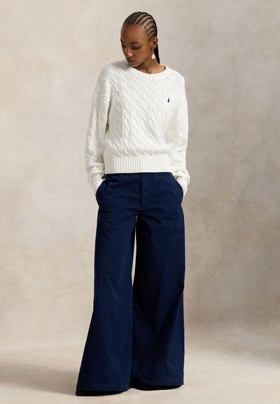 Λευκό πουλόβερ με πλέξη καλωδίου, με ριμπ τελείωμα και μανσέτες, συνδυασμένο με σκούρο μπλε παντελόνι με φαρδιά παντελονοποδιά. Το σύνολο διαθέτει υφή σχεδίασης.