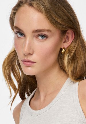 Goudkleurige oorbellen met een golvend ontwerp, gecombineerd met een lichtgrijze mouwloze top. Close-up van het gezicht van een model met natuurlijke make-up.