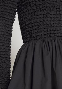 Svart klänning med en texturerad, samlad byst och en flödande kjol. Tyget är slätt med en matt finish och betydande textur.