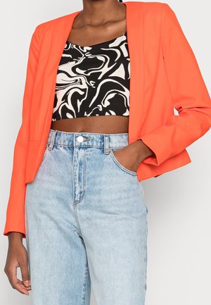 Femme portant un blazer orange vif, un crop top abstrait noir et blanc, et un jean taille haute bleu clair, une main dans la poche.