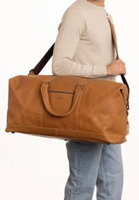 Bolsa de viaje grande de cuero marrón con asas redondeadas, una correa de hombro ajustable y detalles de costura sutiles. Textura suave, diseño espacioso.