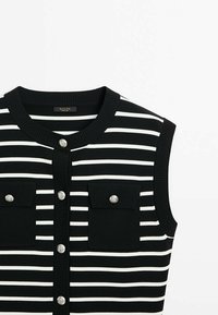 Haut sans manches rayé noir et blanc avec un col rond, deux poches frontales et des accents de boutons métalliques. Tissu côtelé texturé.