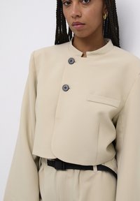 Beige Cropped-Jacke mit Stehkragen, einer Brusttasche und zwei schwarzen Knöpfen. Kombiniert mit hochgeschnittenen Hosen und einem schwarzen Gürtel.
