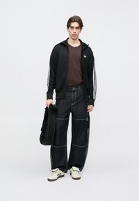 Μαύρο ζακέτα με φερμουάρ και λευκές ρίγες, καφέ μπλουζάκι, φαρδύ σκούρο τζιν παντελόνι και λευκά αθλητικά παπούτσια. Κρατάει μια μαύρη τσάντα.