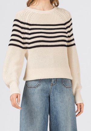 Femme portant un pull en maille crème avec des rayures horizontales noires sur la poitrine et un jean denim bleu clair.