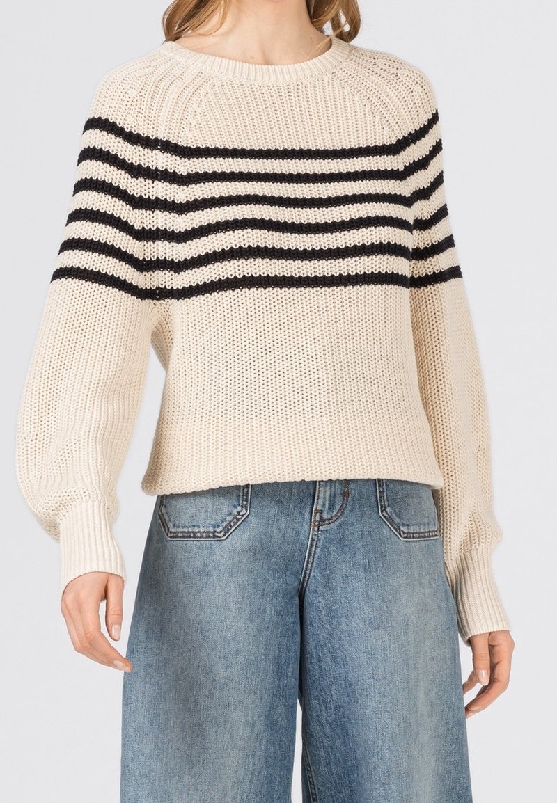 Femme portant un pull en maille crème avec des rayures horizontales noires sur la poitrine et un jean denim bleu clair.