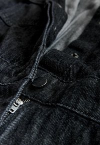 Black denim jacket with a zip closure, silver zipper, rounded collar, and button details. Textured fabric with visible stitching.