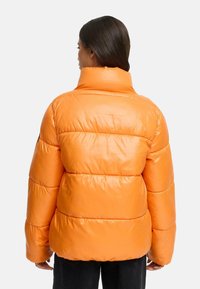 Oranje pufferjack met een hoge kraag, gewatteerd ontwerp en glanzende textuur. De achterkant heeft horizontale naden en een ontspannen pasvorm.